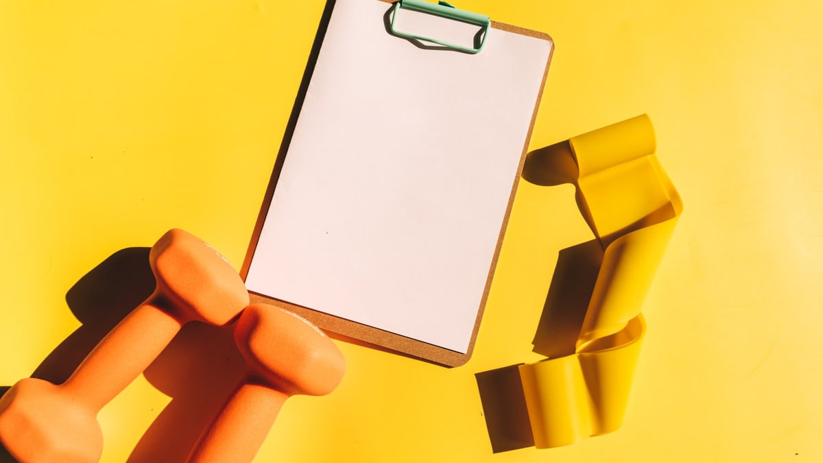 Bright flat lay of orange dumbbells, clipboard, and yellow resistance band on yellow background.